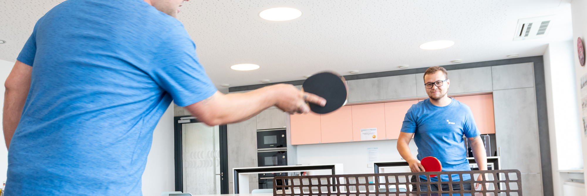 Zwei Mitarbeiter in blauen T-Shirts beim Tischtennis im Aufenthaltsraum